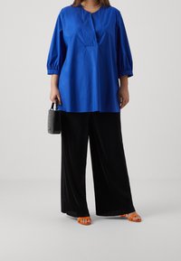 Blå bomullsblus med plisserad halsringning, trekvartslång ärm, tillsammans med svarta vida byxor och orange klacksandaler. Svart väska ingår.