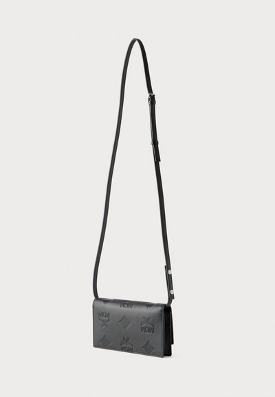 Μαύρη δερμάτινη τσάντα χιαστί με ρυθμιζόμενο ιμάντα, ανάγλυφο λογότυπο MCM και διακοσμητικά μοτίβα σε σχήμα ρόμβου, απεικονίζεται σε απλό λευκό φόντο.