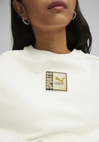 Felpa bianca con girocollo a coste, caratterizzata da un emblema "PUMA" in oro e nero con la parola "SPORTS" e linee radianti dietro di esso.