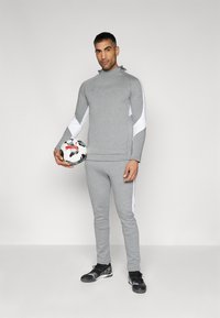 Grå sports tracksuit med hvide accenter, bestående af en hætte-trøje og tætsiddende bukser. Modellen holder en sort og hvid fodbold. Sorte træningssko.