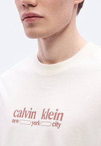 Μπλούζα από βαμβάκι σε κρεμ απόχρωση με στρογγυλή λαιμόκοψη και κοντά μανίκια. Φέρει κόκκινο κείμενο: "calvin klein new york city" ως κύριο γραφικό στοιχείο.