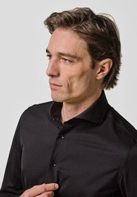 Zwarte longsleeve shirt van gladde stof, met een klassieke kraag en knoopsluiting, gedragen door een persoon met kort haar.