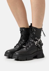 Botas de tobillo de cuero negro con una suela gruesa, cierre de cordones en la parte delantera, correas diagonales con hebillas y una cremallera lateral para facilitar el uso.