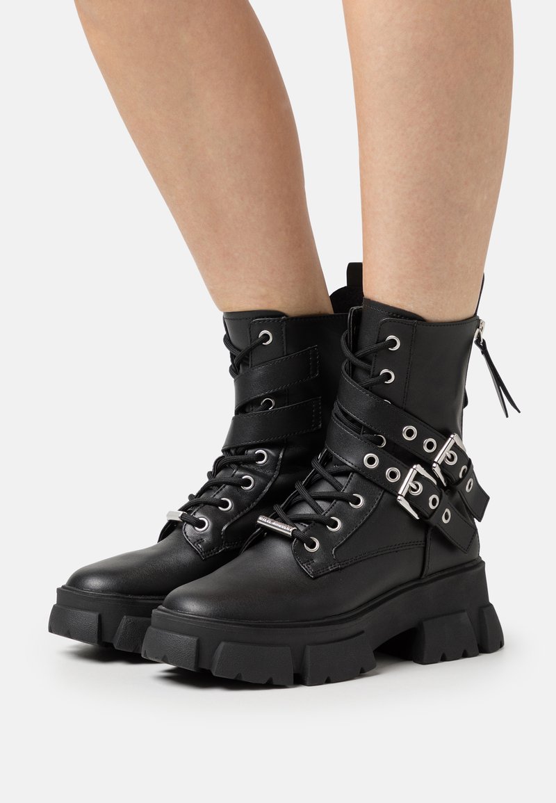 Botas de tobillo de cuero negro con una suela gruesa, cierre de cordones en la parte delantera, correas diagonales con hebillas y una cremallera lateral para facilitar el uso.