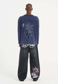 Marineblaues geripptes Sweatshirt mit grafischem Design auf der Rückseite, kombiniert mit lockeren schwarzen Denim-Hosen, die farbenfrohe Tattoo-Elemente aufweisen.