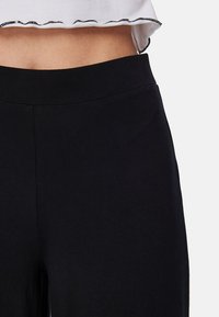 Zwarte broek met een gladde textuur en een elastische tailleband. Heeft een minimale, klassieke uitstraling zonder zichtbare patronen of hardware.