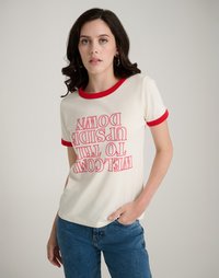 Κρεμ μπλουζάκι με κόκκινες κο�ντές μανίκες και γιακά. Διαθέτει έντονο, αντεστραμμένο κείμενο σε ροζ, που δηλώνει "Καλώς ήρθατε στον Αντεστραμμένο Κόσμο."