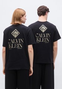 Μαύρα βαμβακερά μπλουζάκια με χρυσό λογότυπο «CALVIN KLEIN» και λουλουδάτο σχέδιο στην πλάτη. Κανονική εφαρμογή με στρογγυλή λαιμόκοψη και κοντά μανίκια.