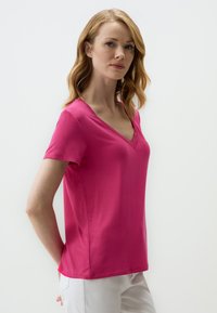 T-shirt rose à manches courtes en V faite d'un tissu lisse ; design ajusté avec une coupe droite et des coutures latérales subtiles, associé à un pantalon blanc.