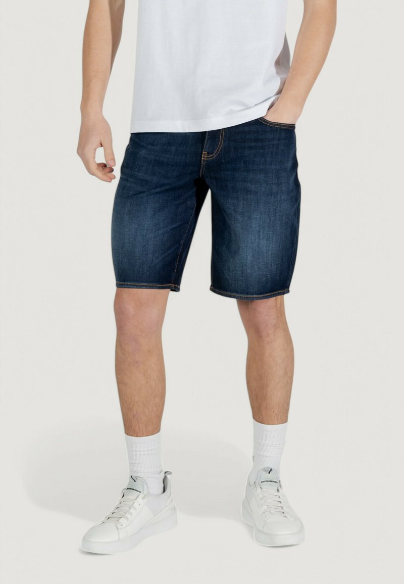 Shorts en denim bleu foncé avec des coutures contrastées, offrant une coupe droite et des poches latérales, associés à des baskets blanches et des chaussettes mi-mollets.