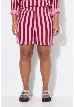 Person iført røde og pink lodrette stribede shorts med matchende skjorte med opkørte ærmer og metallic bukseseler, stående mod en hvid væg.