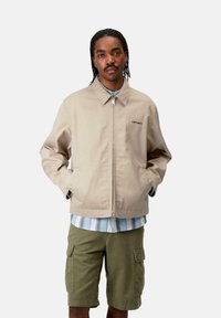 Chaleco beige con cremallera y cuello, con un tejido suave, bolsillos laterales y un logo negro en el pecho, usado sobre una camisa a rayas azul y pantalones cortos verdes.