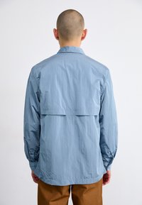 Chemise boutonnée bleu clair en tissu léger, présentant un empiècement au dos et un ourlet arrondi, avec des manches longues et des poignets élastiques.