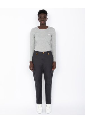 Femme se tenant face à l'avant, portant un t-shirt à manches longues gris clair, un pantalon cargo noir avec des boutons dorés, et des baskets blanches.