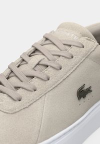Gros plan d'une basket en daim beige avec des lacets assortis, une semelle blanche et un petit logo crocodile foncé sur le côté.