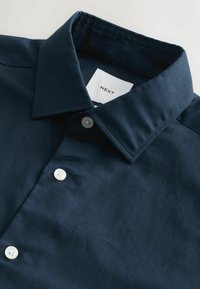 Chemise bleu marine foncé à boutons, col replié et boutons blancs, avec une étiquette intérieure blanche avec texte noir.