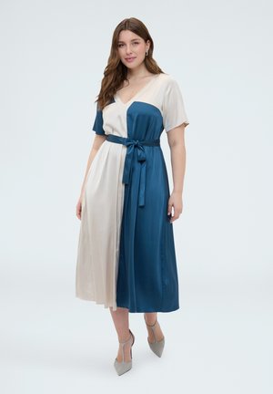 Frau trägt ein midi Kleid in Blau und Creme mit kurzen Ärmeln, V-Ausschnitt, gebundenem Taillengürtel und grauen spitzen Schuhen.