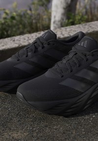 Pair of black Adidas running shoes with laces on outdoor pavement next to a stone curb and green plants in the background.