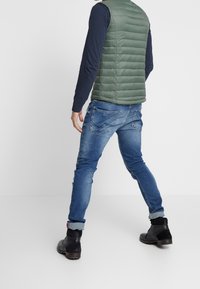 Grön vadderad väst över en marinblå långärmad skjorta, i kombination med blekblå denimjeans och svarta ankelskor. Slät textur med en strömlinjeformad design.
