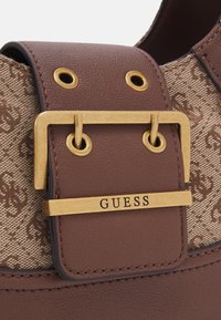 Bolsa de mão em pele castanha com uma fivela dourada com a inscrição "GUESS", acentuada por uma seção em tela padrão clara e ilhós dourados.