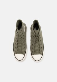 Zapatillas altas de color verde oliva con parte superior de lona, puntera de goma blanca, suela negra y cordones en contraste. Logotipo circular en el lateral.