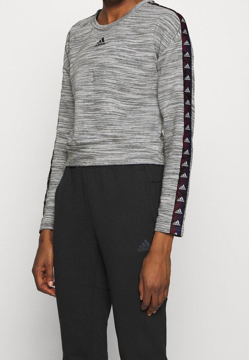 Personne portant un haut gris à manches longues Adidas avec des bandes logo sur les manches et un pantalon noir Adidas avec un logo sur la cuisse gauche.