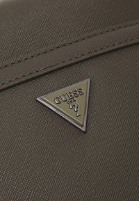 Sac à main gris foncé avec surface texturée, comportant un logo métallique triangulaire en or et en argent avec "GUESS" et un détail de point d'interrogation.