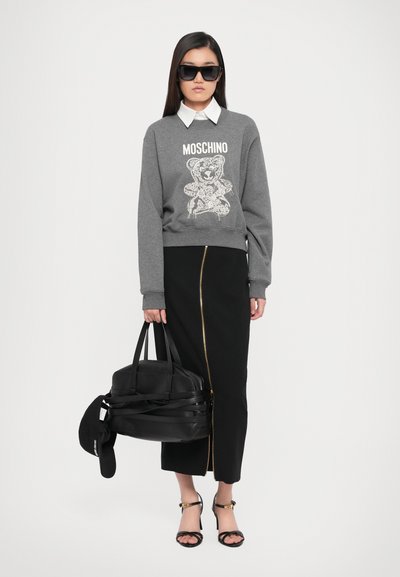 Felpa grigia con grafica di orso e testo "MOSCHINO", gonna midi nera con zip dorata, borsa nera, occhiali da sole e sandali con cinturini.