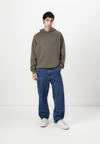 Khaki hooded sweatshirt with a relaxed fit, paired with loose blue denim jeans featuring front pockets and white sneakers.