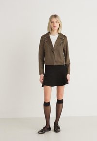 Olijfgroene suède motorjack met zilveren ritsen, gedragen over een wit shirt en een zwarte flared rok, gecombineerd met kniehoge zwarte sokken en ballerina's.