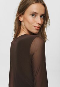 Brun, genomskinlig topp med långa ärmar, mjuk textur, rund halsringning. Modellens hår är vågigt och pryder enkla guldövhängen.