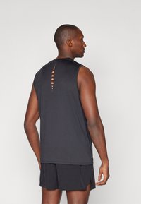 Fekete ujjatlan sporttop, texturált anyagból, kerek nyakkivágással és narancssárga logóval a háton, megegyező nadrággal viselve.