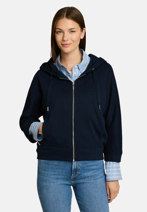 Frau trägt einen dunkelblauen Kapuzenpullover mit Reißverschluss über einem blau-weiß gestreiften Hemd mit Knöpfen und blauen Jeans, steht mit einer Hand in der Tasche.