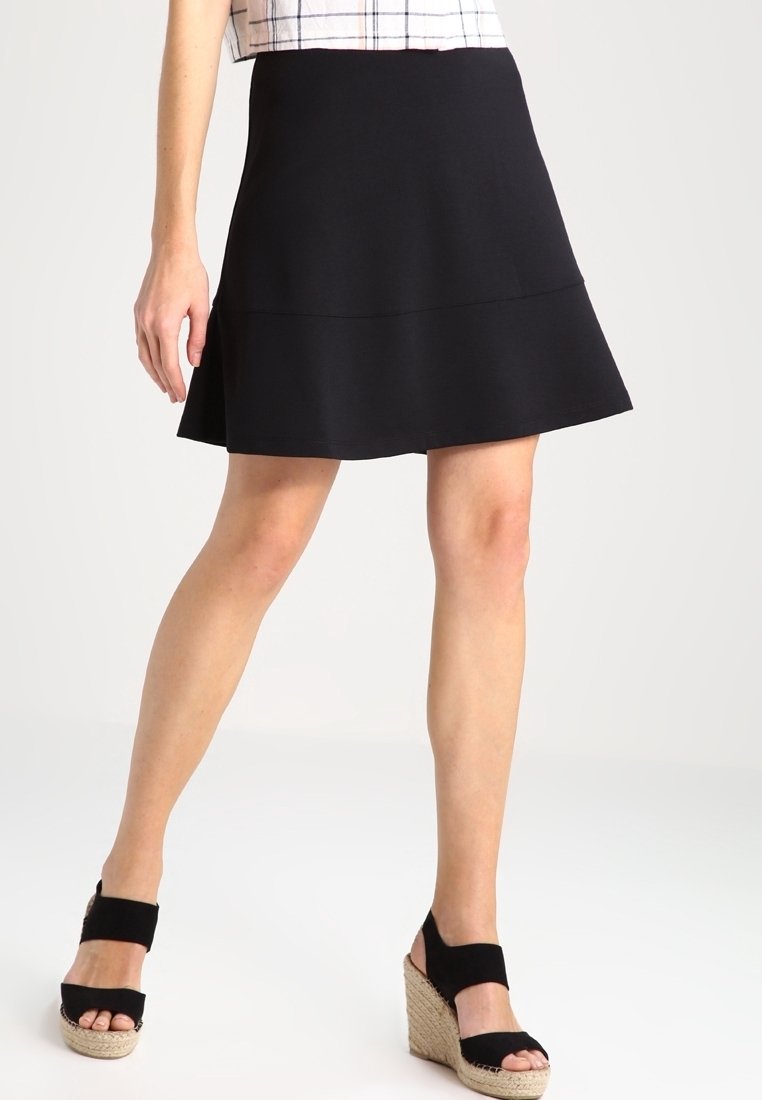 Jupe noire évasée en tissu extensible, avec une texture lisse et une ceinture ajustée. Portée avec des espadrilles noires à talons.