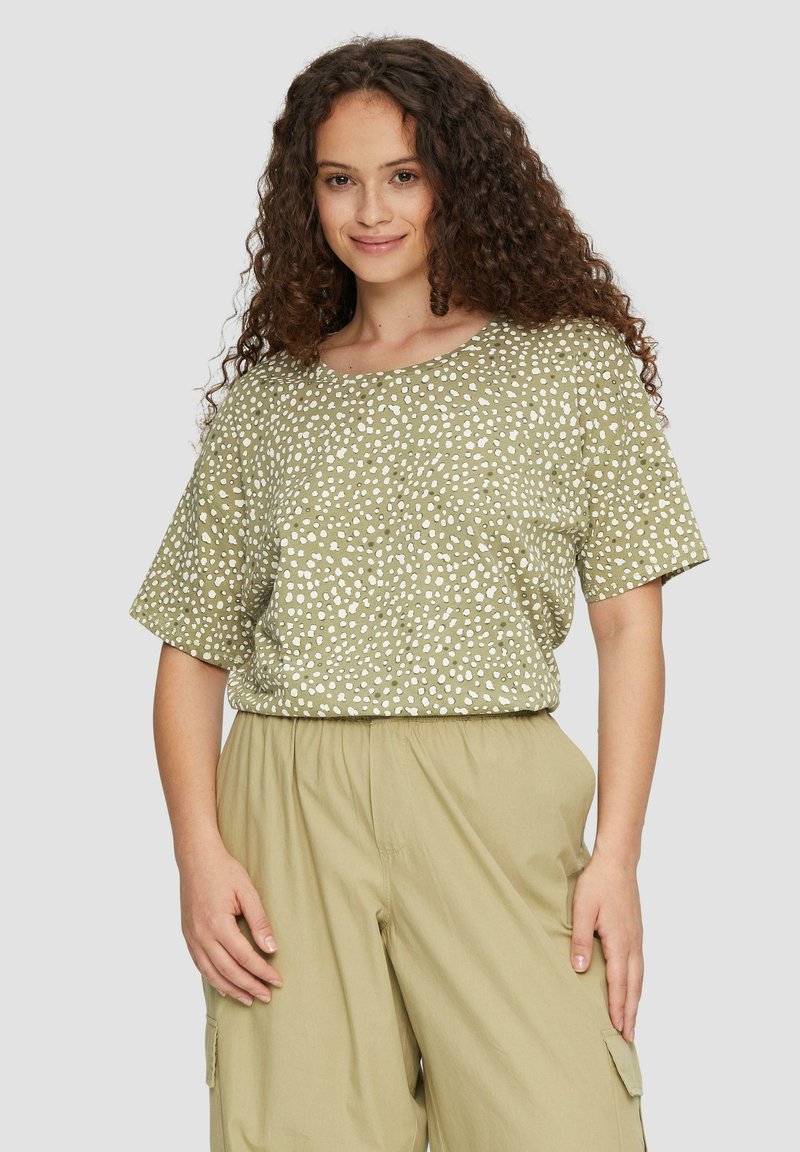 Oliwkowa bluzka z krótkim rękawem w białe kropki, okrągły dekolt i luźny krój, w połączeniu z jasnobeżowymi spodniami.