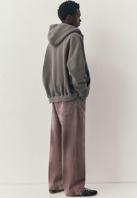 Homme debout, les mains dans les poches, portant un sweat à capuche gris, un jean violet délavé à jambes larges et des baskets noires.