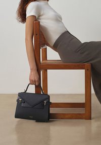 Bolso de mano de piel negra con superficie texturizada, diseño angular, herrajes en tono dorado y un asa superior para llevar.