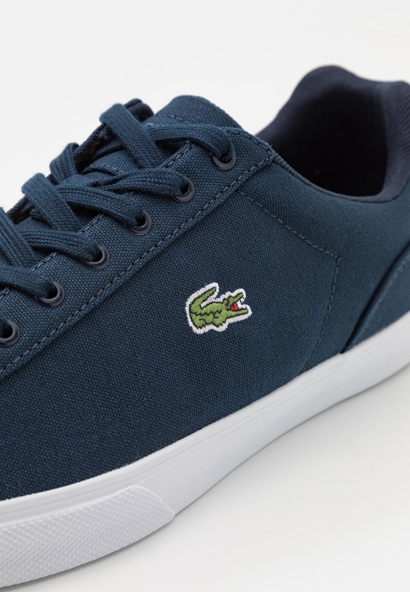 Sneaker di tela navy con suola in gomma bianca, punta rotonda e lacci navy. Presenta un piccolo logo di coccodrillo ricamato di colore verde vicino all'area dei lacci.