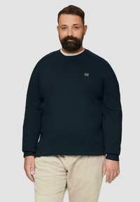 Marineblauwe gebreide trui met een ronde halslijn en ribgebreide mouwen, met een klein logo op de borst. Gecombineerd met lichte corduroy broeken.