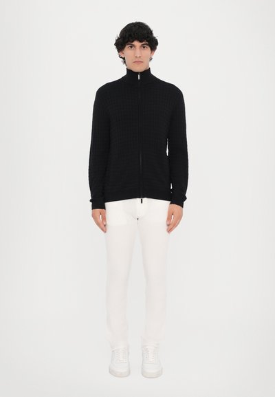 Pulover negru cu fermoar, cu un model texturat în grilă, asortat cu pantaloni albaștri și adidași albi. Design minimalist pe un fundal simplu.