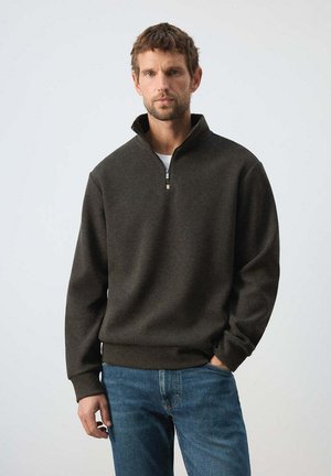 Man met kort bruin haar die een donkergrijze pullover met kwart-rits draagt en een blauwe spijkerbroek, staat met één hand in zijn zak tegen een effen achtergrond.