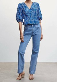 Blusa blu a fantasia con maniche a palloncino, scollatura a V e orlo a peplum, abbinata a jeans dritti azzurri e sandali con tacco trasparente.