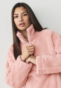 Pile rosa sfocato con cerniera, collo alto e maniche lunghe; tessuto morbido, piccolo dettaglio con cerniera e vestibilità comoda.