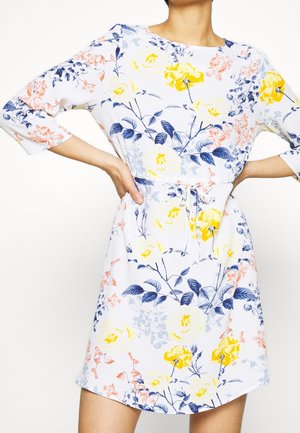 Bílé šaty s žlutým, modrým a růžovým květinovým vzorem, tříčtvrteční rukávy a páskem v pase, nošené osobou s rukama v bok.