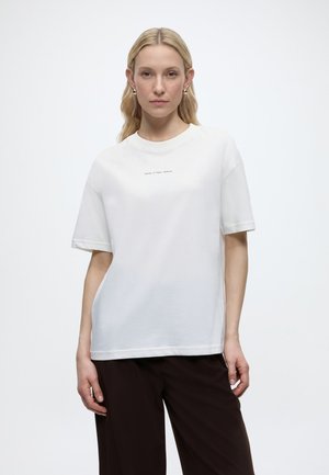 Blondína s okrúhlymi zlatými náušnicami, v voľnej bielej košeli s krátkym rukávom a malým textom uprostred, a v tmavých širokých nohaviciach, stojaca pokojne.