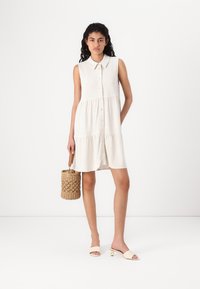 Robe sans manches de couleur beige clair avec un col, un devant à boutons et une jupe évasée. La mannequin porte un sac en osier et des sandales beige à semelle plate.