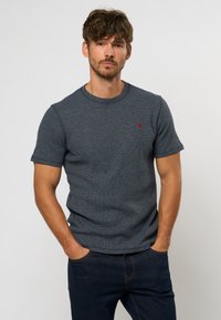 Camiseta de manga corta azul marino con un patrón texturizado y cuello redondo. Presenta un pequeño logotipo rojo en el pecho. Combinada con jeans oscuros.