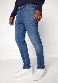 Modré džínové kalhoty s úzkým střihem, klasickým pětipocket designem a jemným vyblednutím, doplněné bílými sportovními teniskami.