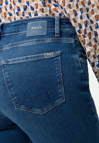 Tmavě modré džíny s obdélníkovou kapsou a značkovou nášivkou na pase. Látka má jemnou texturu a mírné vyblednutí.