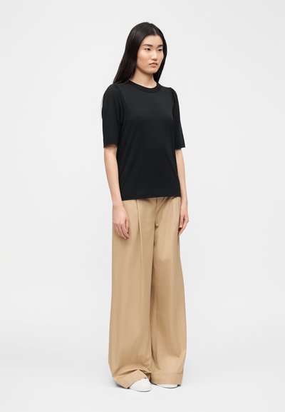 Femme aux longs cheveux bruns portant un haut noir à manches courtes, un pantalon beige large et des chaussures blanches, debout devant un fond clair uni.